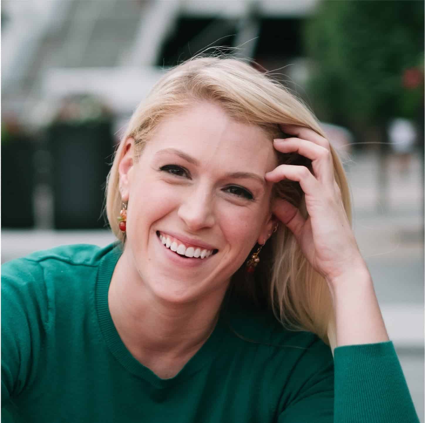 A smiling woman with blond hair wearing a green top and earrings, with her hand in her hair.