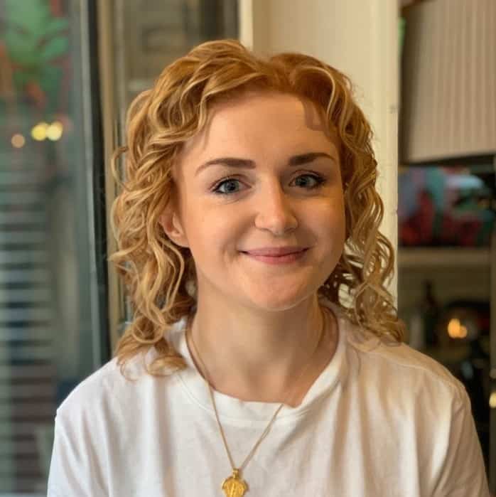 A woman with curly blonde hair and a white t-shirt smiling at the camera.