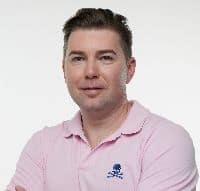 A person with short hair wears a pink polo shirt with a blue emblem. The background is plain white.