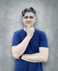 A person with short hair stands against a gray textured wall, wearing a blue t-shirt. They have one hand under their chin and the other arm crossed, with a thoughtful expression.