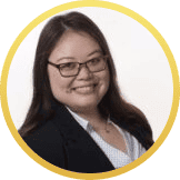 A woman with long dark hair and glasses smiles at the camera. She is wearing a black blazer over a white shirt and is set against a plain white background with a gold circular border.