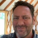 A man with short hair and a beard is smiling slightly. He is indoors with wood beams and a window in the background.