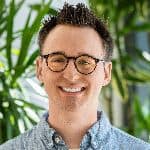 A person wearing glasses and a blue shirt smiles in front of green plants.