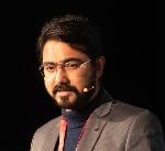Man wearing glasses and a gray suit speaks into a microphone headset against a dark background.