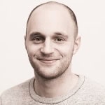 A person with a shaved head and beard is wearing a light sweater and smiling at the camera.