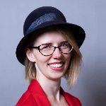 Person wearing a black hat and glasses, smiling, and dressed in a red outfit against a gray background.