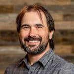 A person with a beard and long hair smiles, wearing a grey shirt. A wooden wall background is visible.