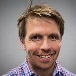 Smiling person with short hair, wearing a checkered shirt, against a plain gray background.