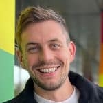 Smiling person with short hair and a beard, standing indoors against a colorful background.