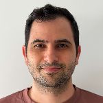 A person with short dark hair and a slight smile is wearing a pinkish shirt against a plain white background.