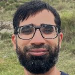 A person with a beard and glasses is outdoors, with grass in the background.