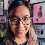 A person with glasses smiles at the camera, with framed pictures on the wall in the background.