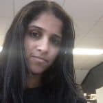 Person with long dark hair in an indoor setting, looking at the camera. Fluorescent lights are visible in the background.