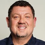 A person with short curly hair and a goatee smiles, wearing a dark shirt against a plain background.