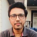 A person with glasses, facial hair, and short dark hair is looking at the camera, seated outdoors with a building in the background.