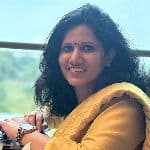 Woman in a yellow outfit smiling while sitting indoors near a window with a blurred green landscape in the background.