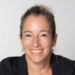 A smiling person with light skin and brown hair is wearing a black blazer against a gray background.