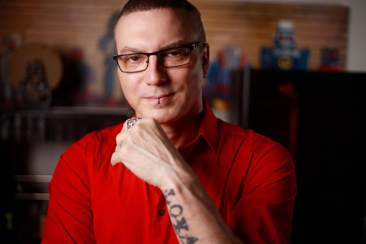 A person wearing glasses and a red shirt is sitting, resting their chin on their hand. A tattoo is visible on their forearm—perhaps symbolizing the seamless fusion of creativity and automation that defines the AI era.