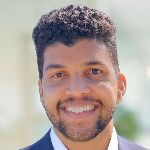 Smiling man with short curly hair and a beard, wearing a suit, looking at the camera.