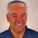 A smiling man with gray hair wearing a blue shirt against a plain background.