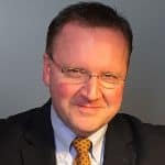 Man in glasses, wearing a dark suit and patterned tie, smiling slightly with a neutral background.