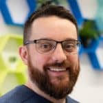 A person with glasses and a beard smiles at the camera, standing indoors with a colorful geometric background.