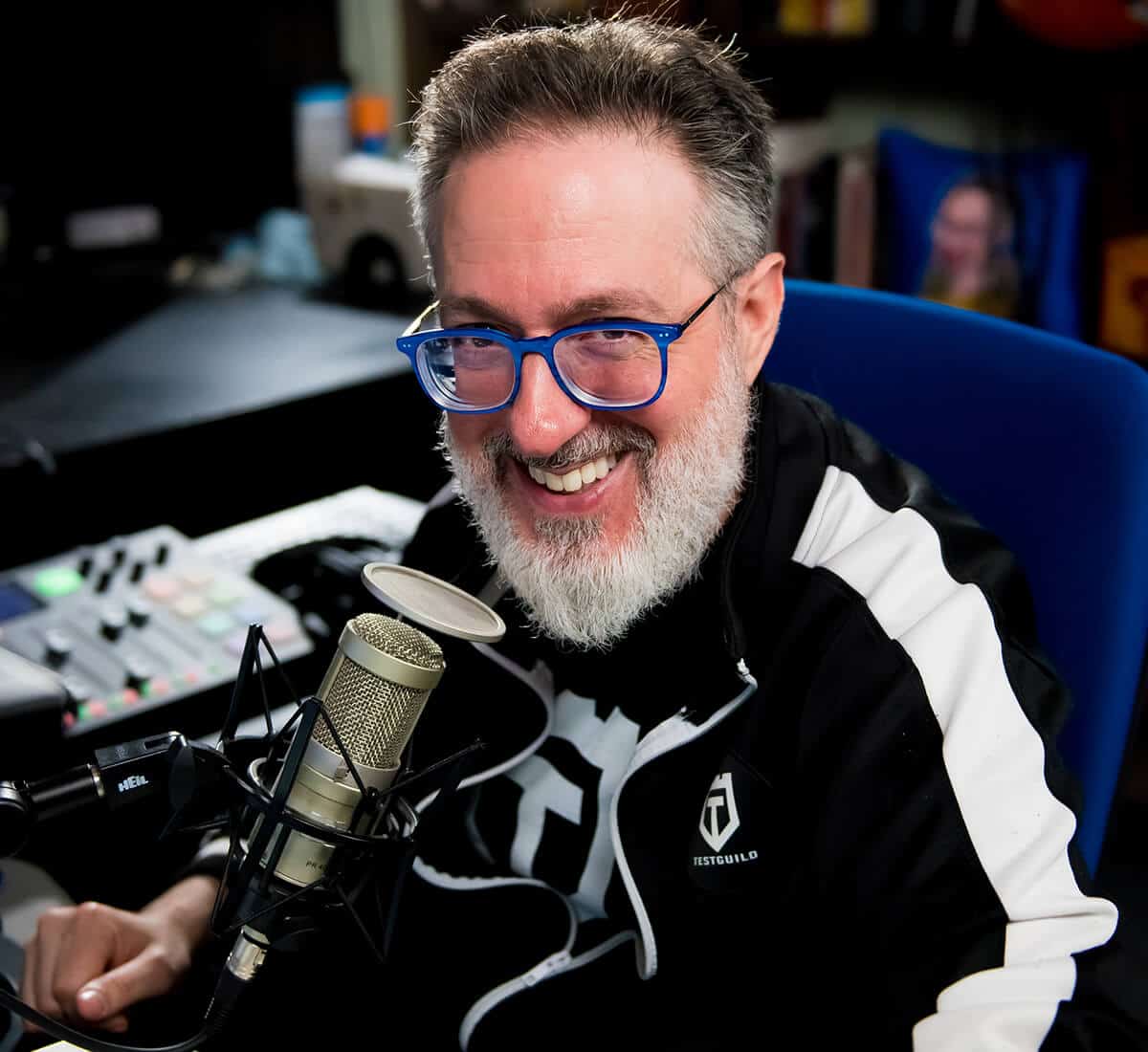 A bearded man with blue glasses and a black-and-white jacket smiles at a microphone in a studio setting.