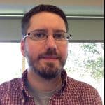 A person with glasses, beard, and short hair, wearing a red checked shirt over a gray t-shirt, is sitting indoors in front of a window with trees visible outside.