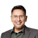 Smiling man wearing glasses, a gray blazer, and a dark green shirt against a white background. Smiling man wearing glasses, a gray blazer, and a dark green shirt against a white background.