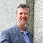 A man with short gray hair and a beard, wearing a blue blazer and light blue shirt, stands and smiles in front of a gray stone wall. A man with short gray hair and a beard, wearing a blue blazer and light blue shirt, stands and smiles in front of a gray stone wall.