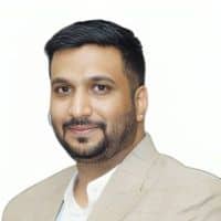 A man with short dark hair and a beard, wearing a light-colored suit jacket and white shirt, poses against a plain white background.