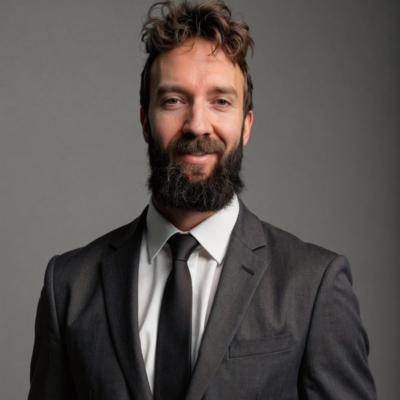 A man with a full beard and tousled hair, wearing a dark suit, white shirt, and black tie, stands in front of a plain gray background, smiling slightly.