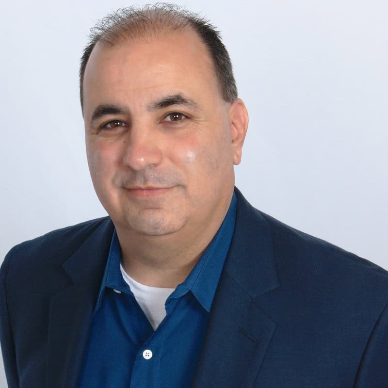A middle-aged man with short hair, wearing a dark blazer over a blue shirt, poses confidently in front of a plain light background—embodying professionalism and quality in the fast-paced AI era.
