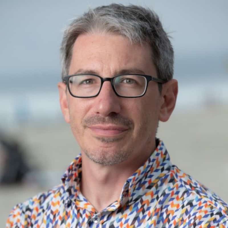 A man with short gray hair, glasses, and a patterned shirt stands outdoors with a blurred beach background, as if pondering innovations in AI Agents.