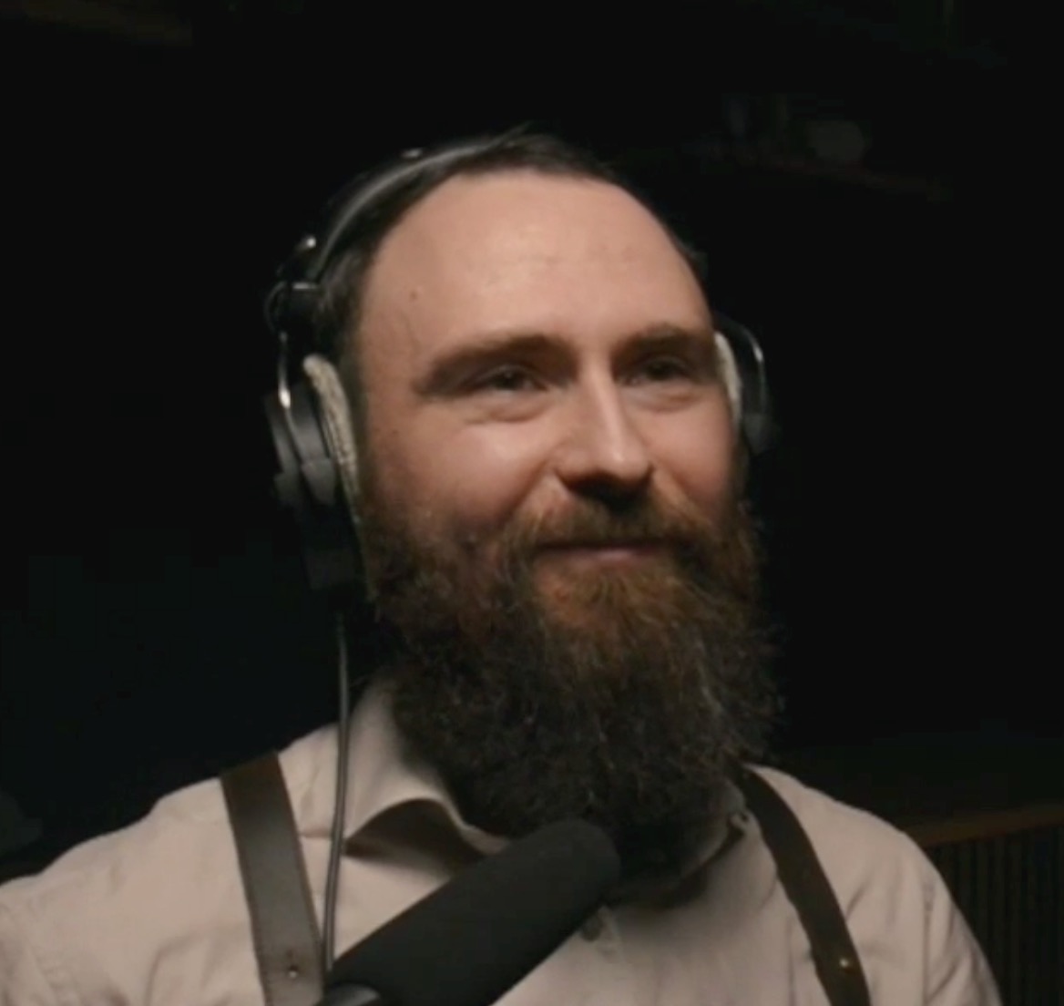 Anton Malinski, sporting a full beard and headphones, smiles while discussing Testing Pipelines into a microphone, dressed in a light-colored shirt and suspenders against a dark background.