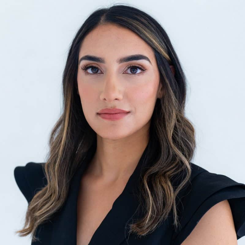 Jennifer Rahmani, with long, wavy brown hair and a black top, looks directly at the camera against a plain light background, embodying confidence and focus—much like Kubernetes enables AI-Powered Predictive Autoscaling in modern tech environments.