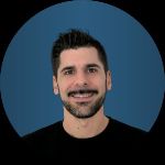 A man with short dark hair and a beard, wearing a black shirt, smiles in front of a blue circular background.