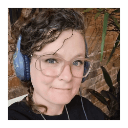 A person with curly hair, glasses, and blue headphones smiles slightly while sitting indoors in front of a brick wall and plants.