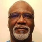 A man with short gray hair, a full gray beard, and glasses, wearing a blue shirt, is facing the camera against a plain light background.