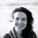 A woman with curly dark hair smiles at the camera in an outdoor setting, with a blurred background. The photo is in black and white.