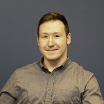 A person with short brown hair in a gray button-up shirt sits and smiles in front of a plain dark blue background.
