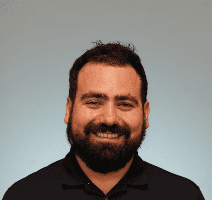 Wilhelm Haaker, a man with dark hair and a full beard, wearing a dark polo shirt, smiles at the camera against a plain light blue background.
