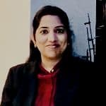 A woman with dark hair is smiling, wearing a dark blazer over a red top, and sitting indoors with a neutral background.