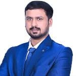 A man with short dark hair and a beard, wearing a blue suit, blue tie, and white shirt, poses with arms crossed against a white background.