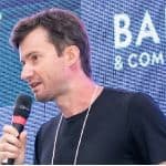 A man with short brown hair speaks into a microphone in front of a blue screen displaying partial text.