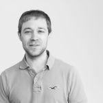A man with short hair and light facial hair is wearing a light-colored polo shirt, standing in front of a plain white background.