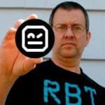 A person holding a round black sticker with a white winking face icon, standing outdoors in front of a white wall.