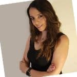 A woman with long brown hair, wearing a black sleeveless top and a watch, stands with arms crossed, smiling slightly in front of a neutral background.