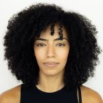 A woman with medium-dark skin and natural, curly black hair looks straight at the camera with a neutral expression. She is wearing a sleeveless black top.