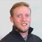 A man with short blond hair and a beard, wearing a dark zip-up sweater, smiling at the camera against a light background. A man with short blond hair and a beard, wearing a dark zip-up sweater, smiling at the camera against a light background.